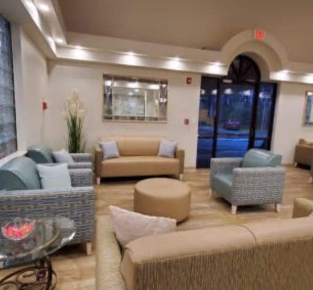 A well-lit living room area with beige and blue cushioned sofas and armchairs arranged around a round beige ottoman. There is a glass side table with a decorative bowl, a large mirror on the wall, and a tall plant in the corner. The room has wooden flooring and a large arched glass door leading outside.