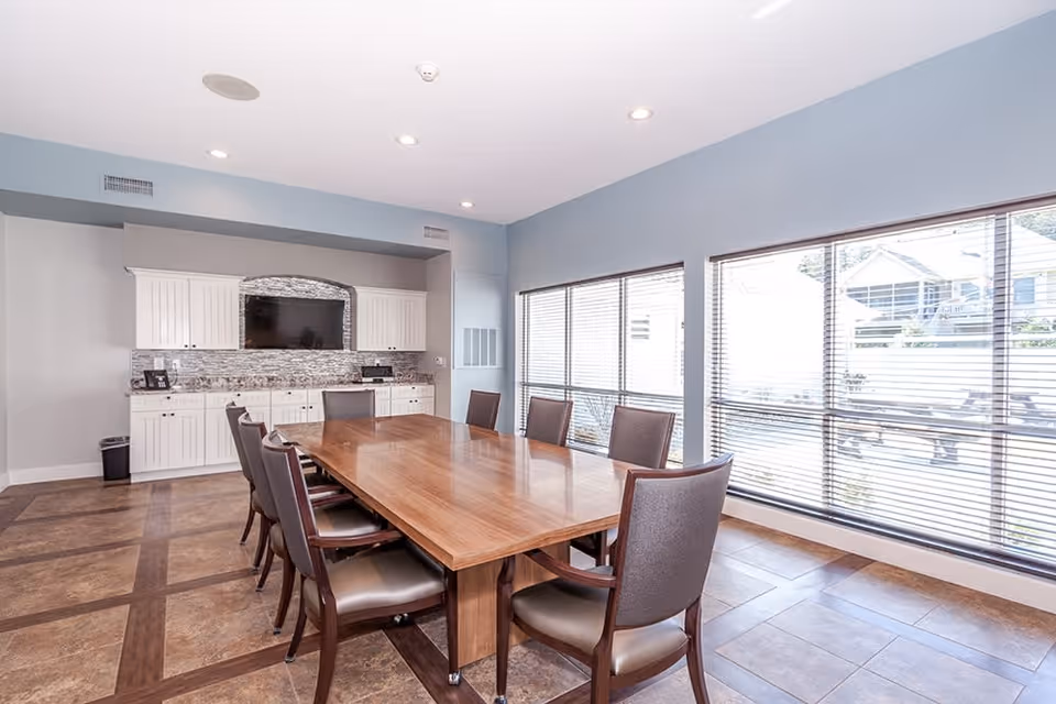 A bright dining room with a large wooden table surrounded by eight chairs. The room has large windows with blinds letting in natural light, and white cabinetry with a mounted flat-screen TV on the far wall. The floor is tiled with a wood inlay pattern.