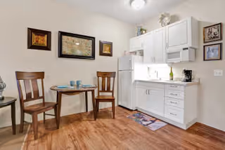 Small kitchen area with white cabinets, a white refrigerator, and a countertop with a coffee maker. Adjacent to the kitchen is a small round wooden table set with two place settings and two wooden chairs. The walls have framed artwork, and the floor is wood laminate.