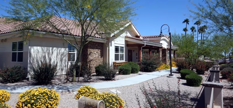 Single-story stucco and stone memory care building with a walkway, desert landscaping, and a lamppost under a clear blue sky.