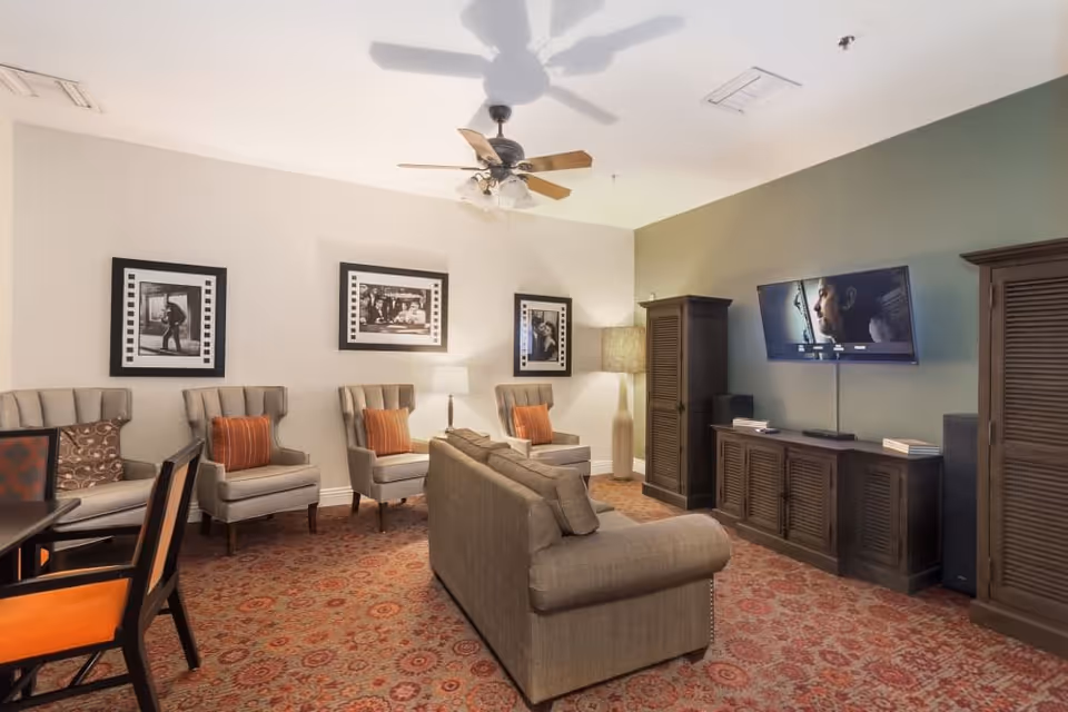 A cozy living room with a beige sofa and three armchairs with orange and patterned cushions. There is a wooden dining table with chairs on the left side. The room has a patterned carpet, a ceiling fan with lights, and three black-and-white framed photographs on the wall. A flat-screen TV is mounted on a green accent wall above a wooden cabinet, flanked by two tall wooden storage units. A floor lamp stands in the corner.