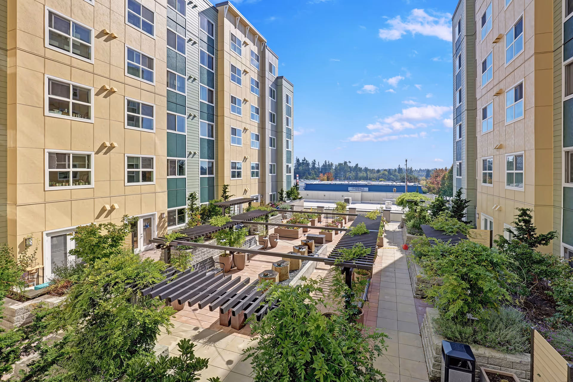 Outdoor courtyard area between two multi-story residential buildings with beige and green exterior walls. The courtyard features paved walkways, raised garden beds with lush greenery, wooden pergolas, and seating areas. The sky is clear with a few clouds.