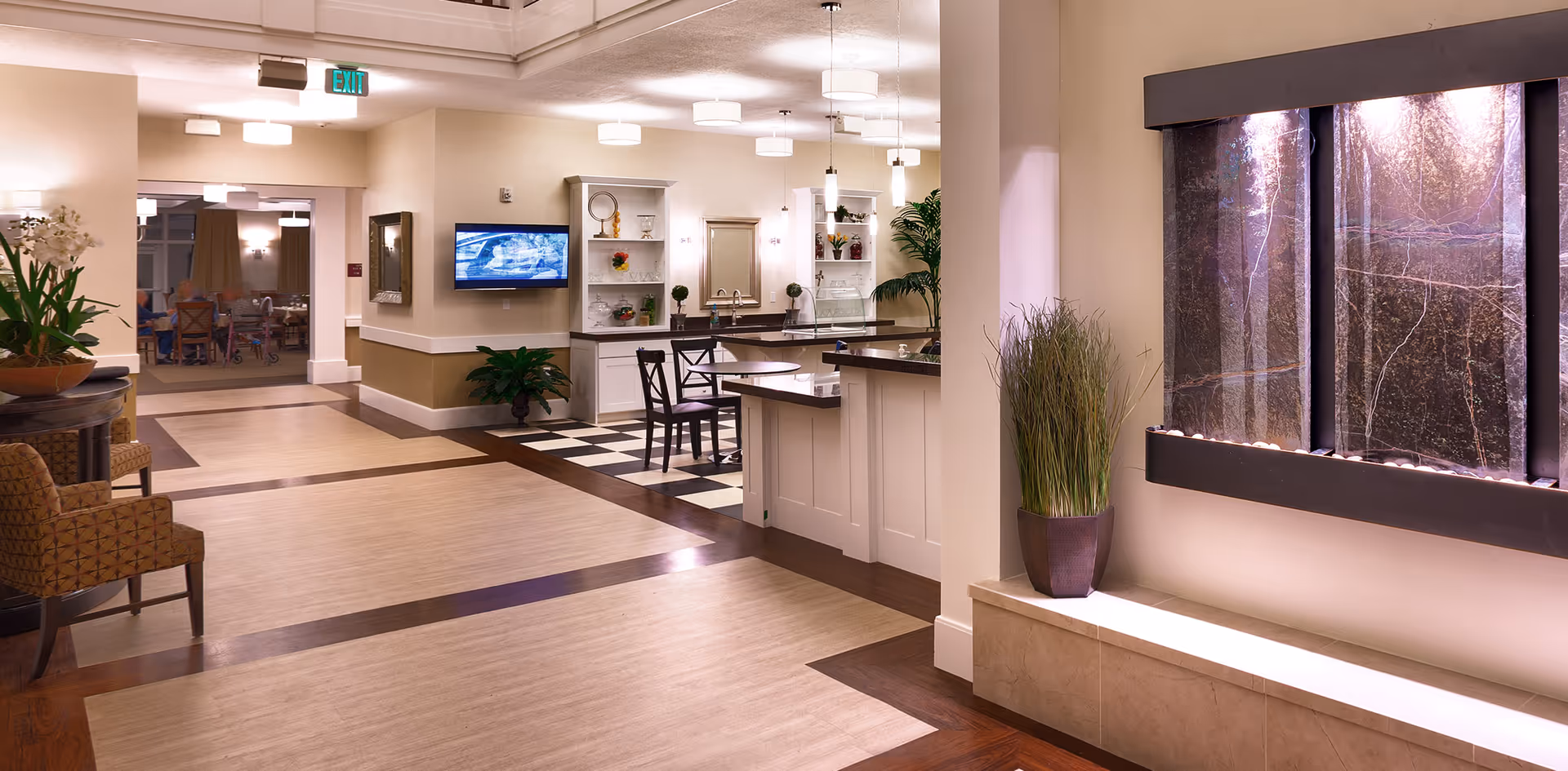 Interior view of a senior living facility hallway with a seating area, a small dining nook with tables and chairs, a mounted TV, decorative plants, and a water feature on the wall.
