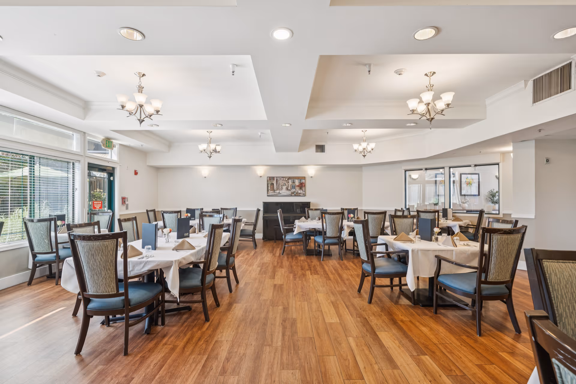 A bright and spacious dining room with multiple tables covered in white tablecloths, each set with napkins, menus, and small flower arrangements. The room features wooden flooring, large windows with blinds allowing natural light, and elegant chandeliers hanging from the ceiling.
