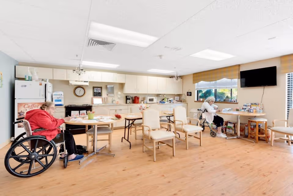 Communal dining/activity room with a kitchenette, tables and chairs, and two elderly residents seated (one in a wheelchair).