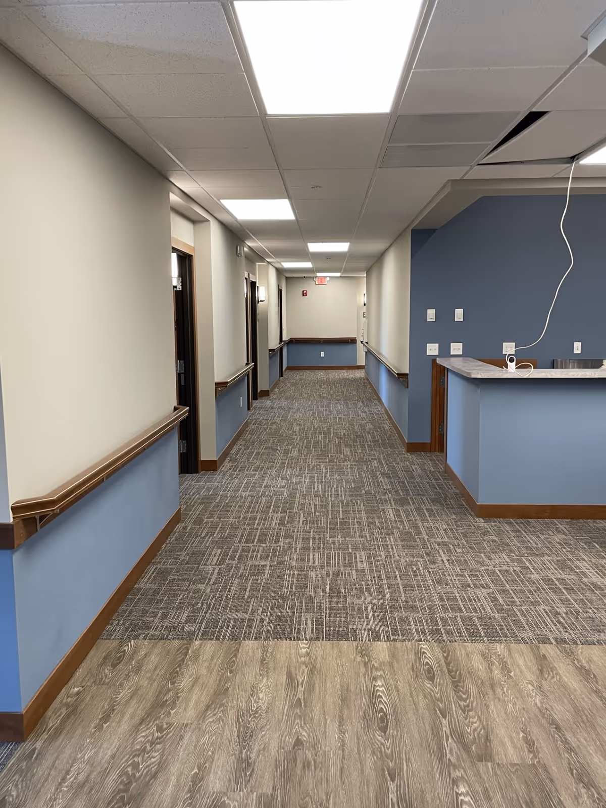 Carpeted interior hallway of a senior living facility with handrails, doorways along both sides, and a reception counter on the right.