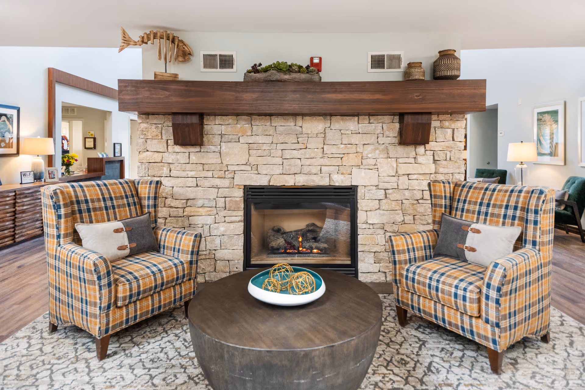 A cozy living room area featuring a stone fireplace with a wooden mantel. Two plaid armchairs with decorative pillows are positioned on either side of a round wooden coffee table with a decorative bowl containing woven spheres. The room has light-colored walls, wooden flooring, and additional seating and lamps visible in the background.