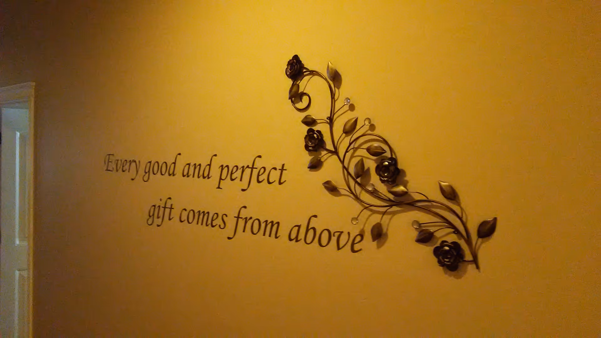 A warm-toned interior wall decorated with a metal floral wall sculpture and the quote "Every good and perfect gift comes from above."
