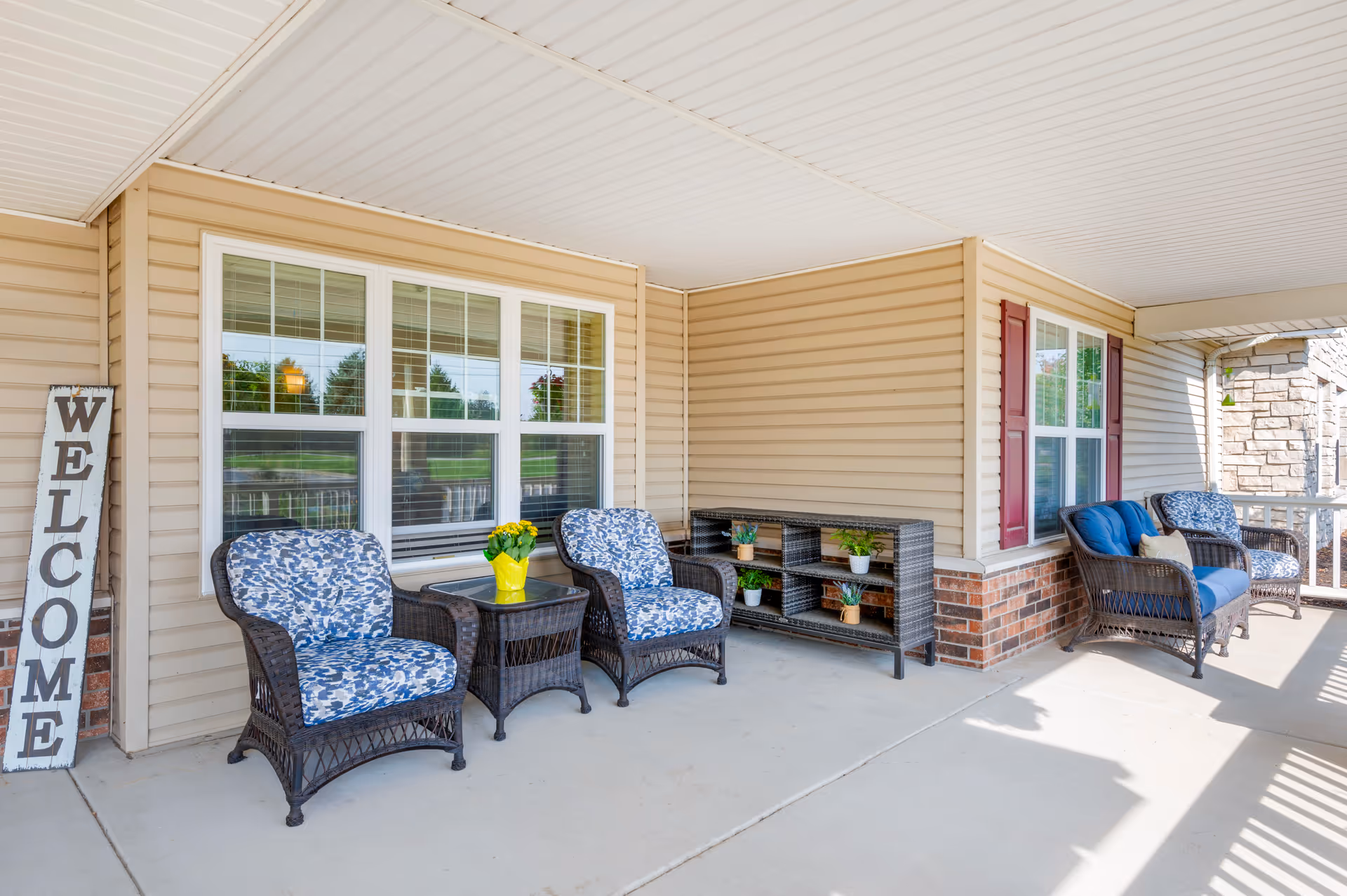 Covered front porch with wicker chairs, small tables, potted plants and a 'WELCOME' sign.