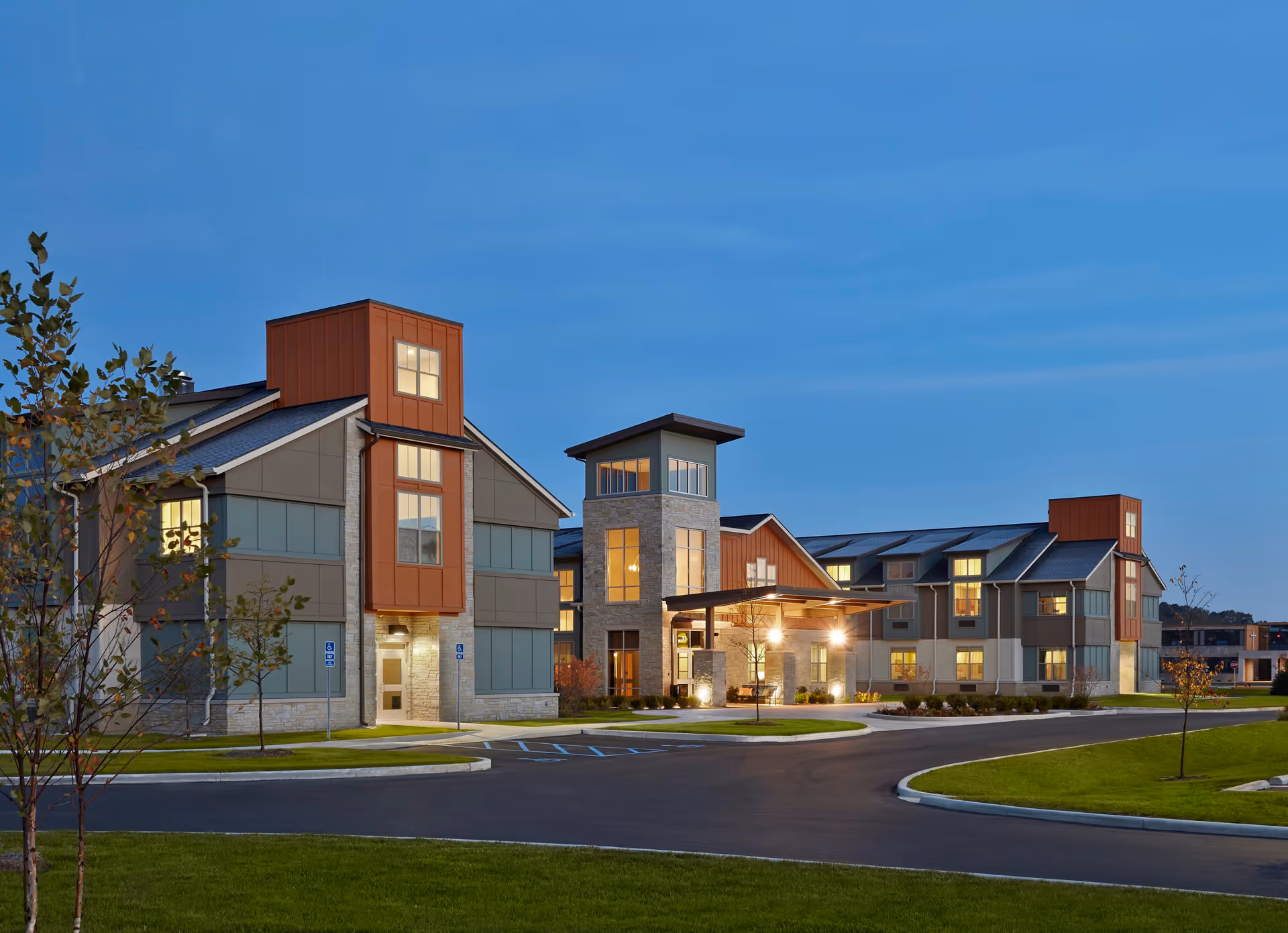 Exterior view of Wellbrooke of South Bend facility at dusk, showing a modern multi-story building with large windows, stone and metal accents, a covered entrance, surrounding landscaping, and a curved driveway with parking spaces.