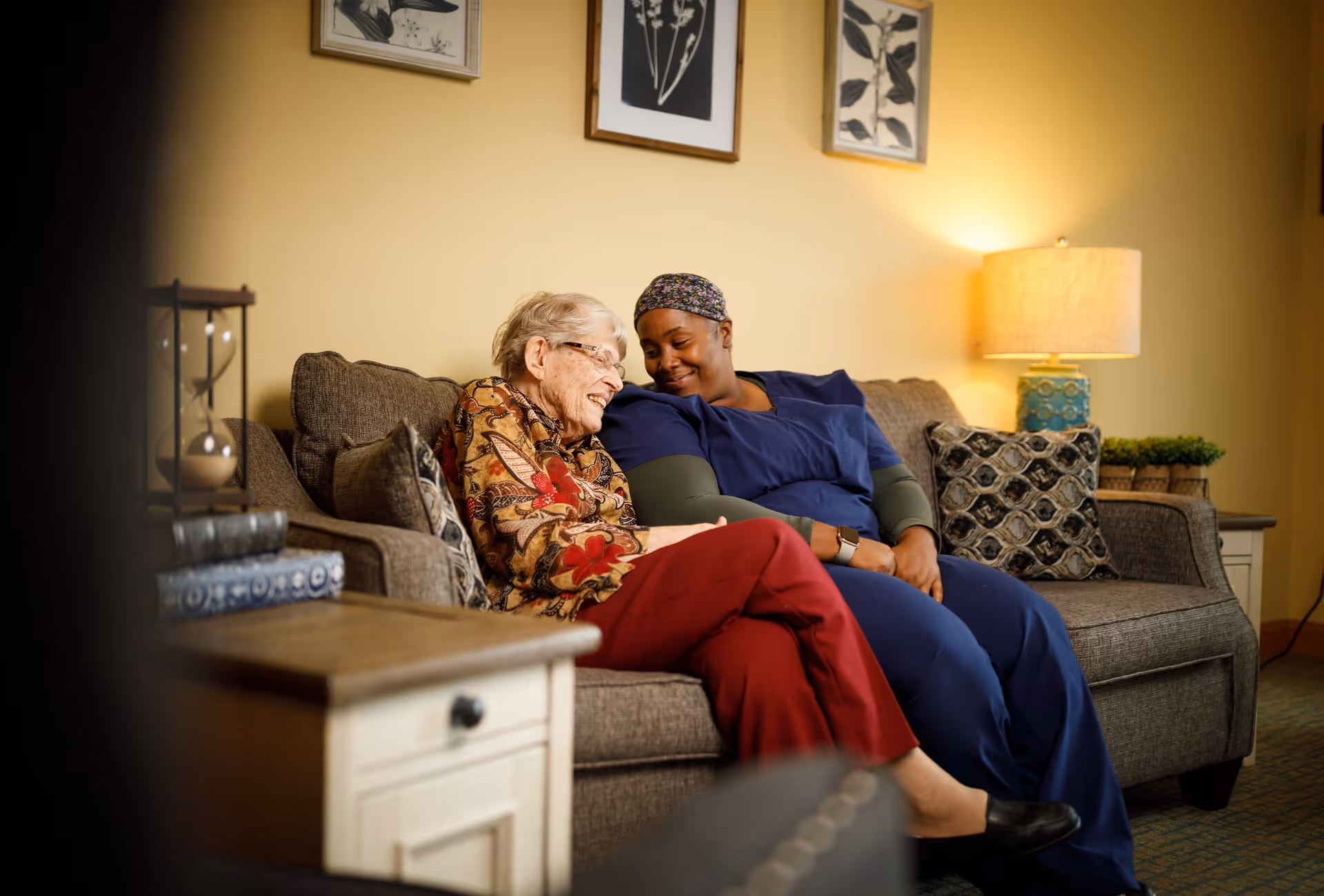 An elderly woman and a caregiver sitting closely together on a couch in a warmly lit living room, sharing a tender moment and smiling at each other. The room has framed botanical artwork on the wall, a side table with an hourglass and books, and a lamp providing soft light.