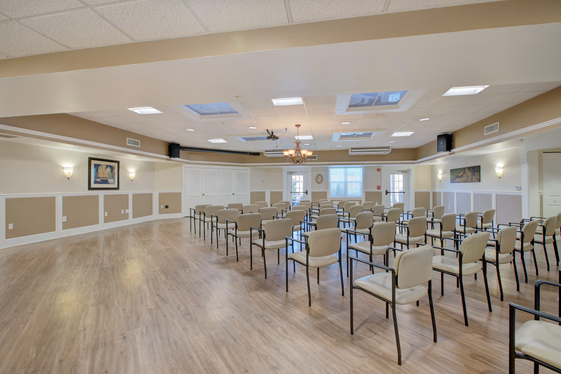 A spacious meeting or activity room with rows of beige chairs arranged facing the front. The room has light wood flooring, beige and white walls with framed artwork, ceiling lights, and skylights. There is a chandelier hanging from the ceiling and windows at the front letting in natural light.
