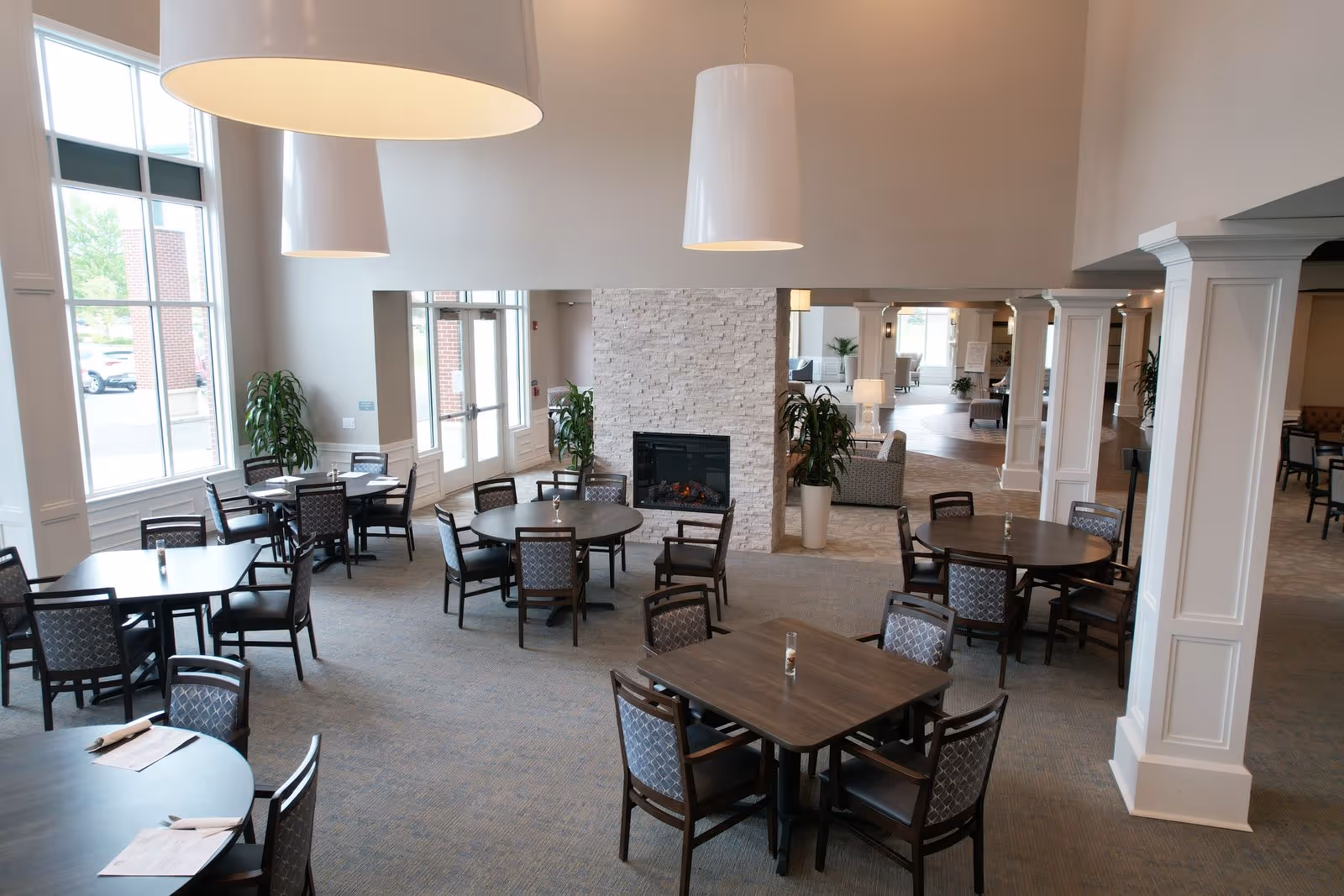 Open dining room with multiple tables and chairs, large windows, hanging lights, and a central stone fireplace.