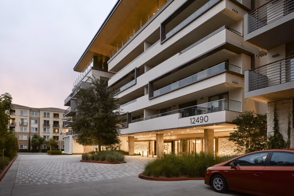 Exterior view of a modern multi-story residential building with balconies, a driveway, landscaped greenery, and a red car parked in front. The building number 12490 is visible above the entrance.