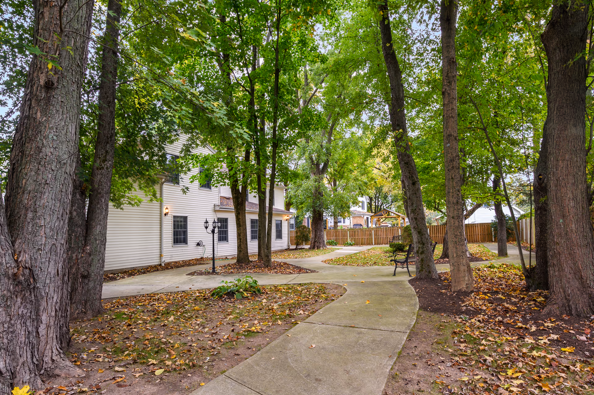 A peaceful outdoor garden area at Fox Trail Memory Care Living with a winding concrete pathway surrounded by tall trees with green leaves. There is a white two-story building on the left side, a black street lamp, a bench along the path, and a wooden fence enclosing the area in the background.