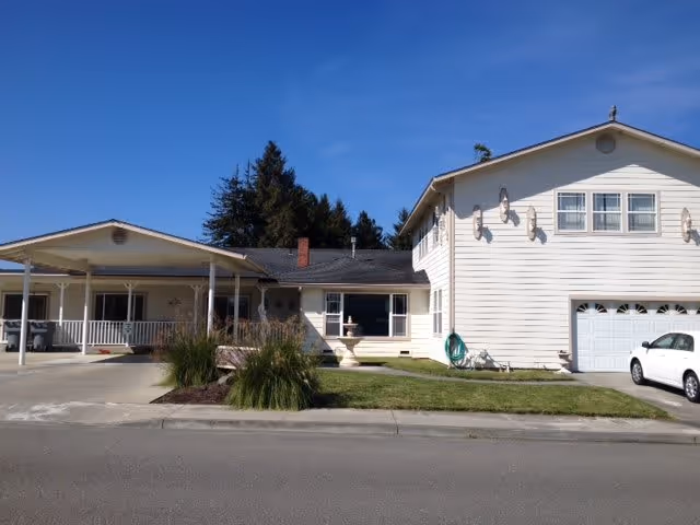 Front exterior of a two-story light-colored residential building with a covered porch, garage, driveway, small lawn, and a white car under a clear blue sky.