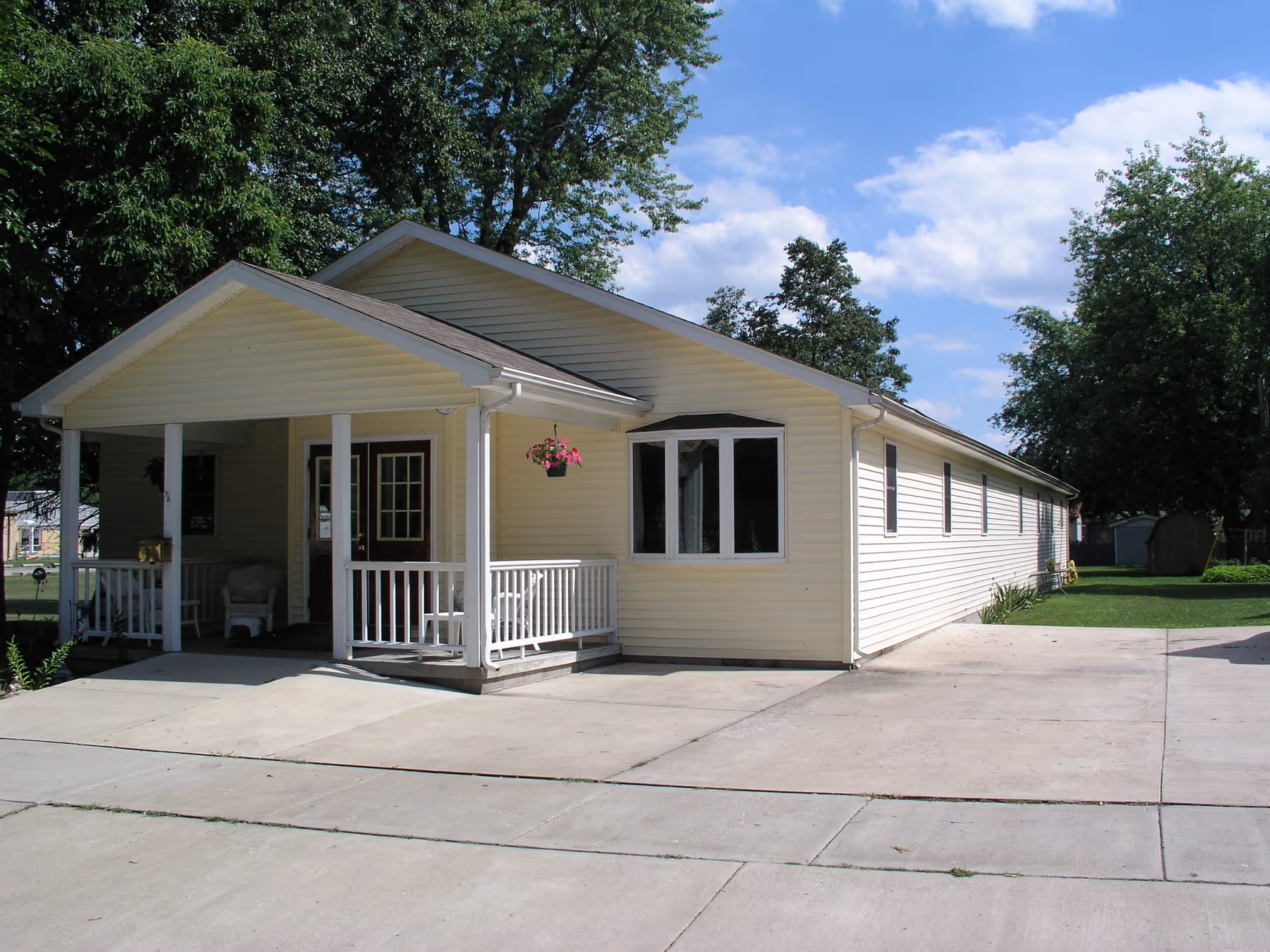 Single-story light yellow building with a covered porch, white railings, and a concrete driveway. There are trees and a grassy area surrounding the building under a partly cloudy blue sky.