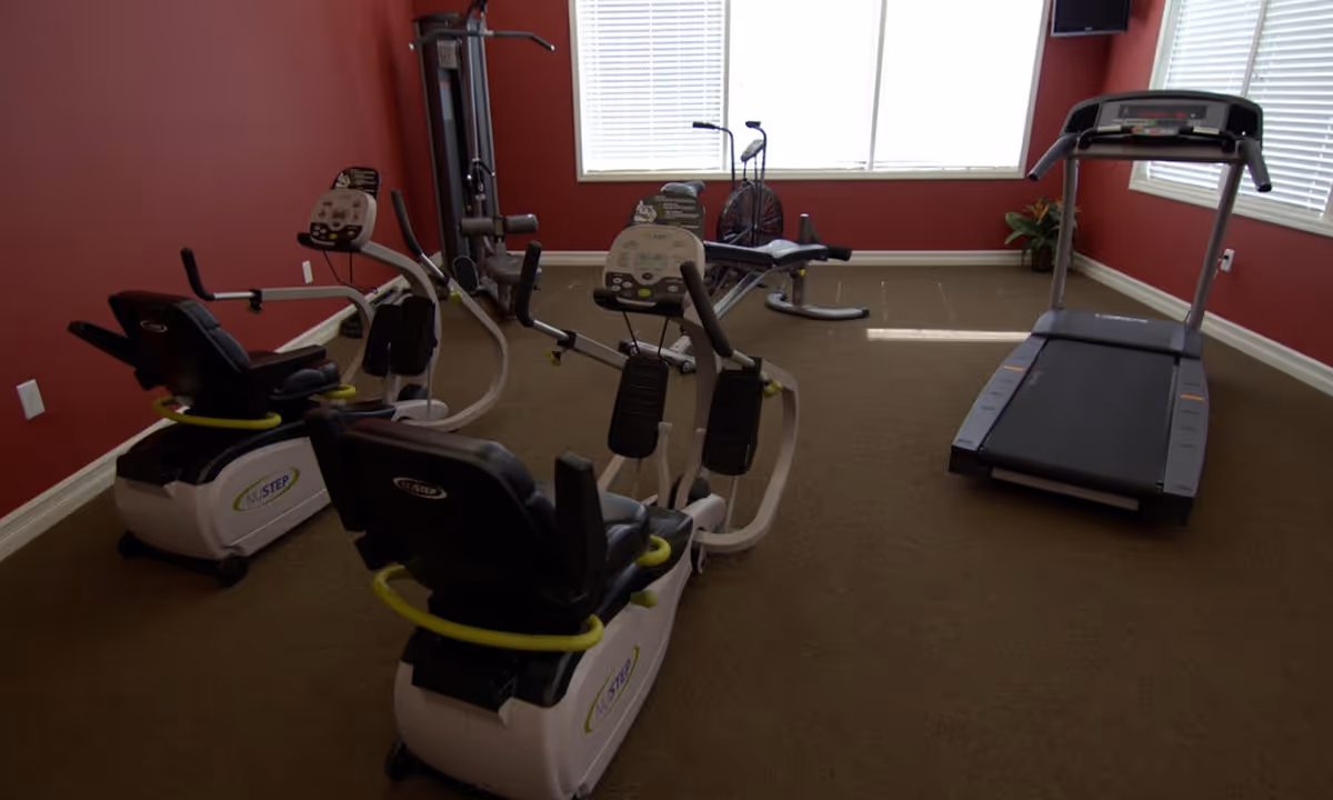 Exercise room with two NuStep recumbent cross trainers, a treadmill, and a multi-station weight machine against red walls and carpeted floor, with windows letting in natural light.