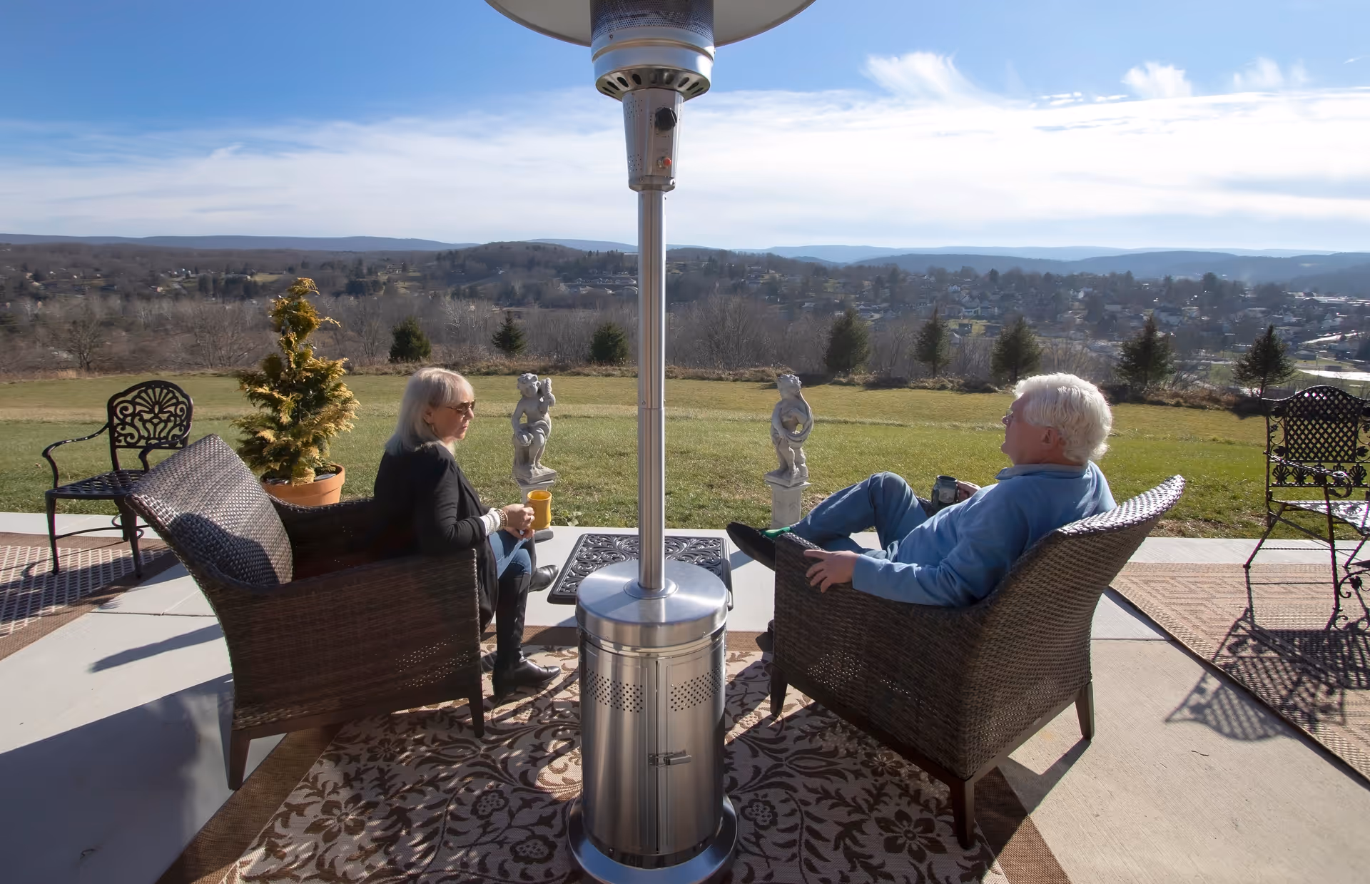 Two elderly people sitting on wicker chairs on a patio with a tall outdoor heater between them, overlooking a grassy area and a distant view of hills and houses under a partly cloudy sky.