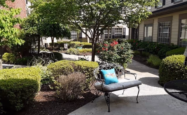 Outdoor garden area at The Carriages of Rivergate featuring a paved walkway, manicured bushes, flowering plants, a tree, and a metal bench with a cushion and decorative pillow. The building with multiple windows is visible in the background.