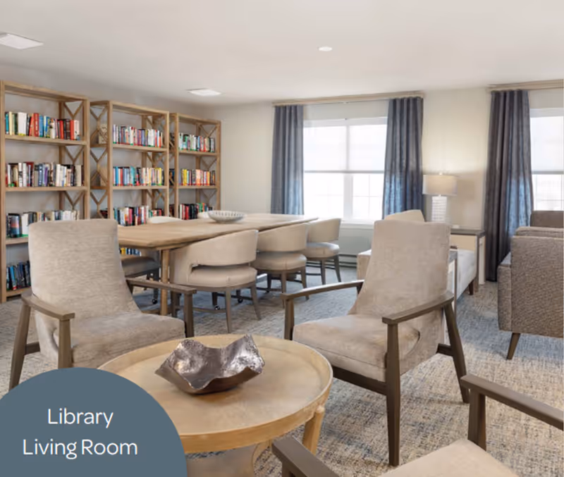 A bright and cozy library living room featuring a wooden table surrounded by beige cushioned chairs, bookshelves filled with books along the wall, large windows with blue curtains, and a small round wooden coffee table with a decorative bowl on it.