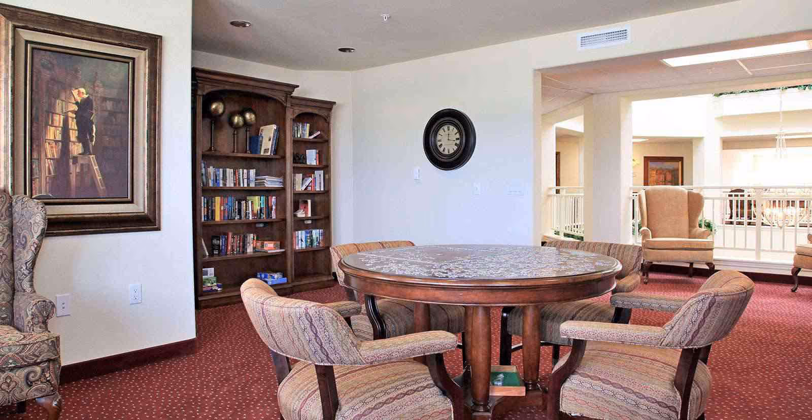 A senior living common area with a round wooden game table surrounded by upholstered chairs, a bookshelf, and armchairs overlooking an open atrium.