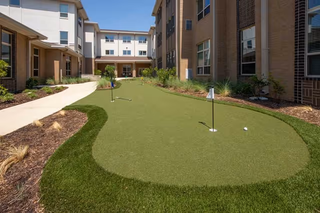 Outdoor putting green with three golf holes and flags situated between two buildings under a clear blue sky, with landscaped plants and a paved walkway on the left side.