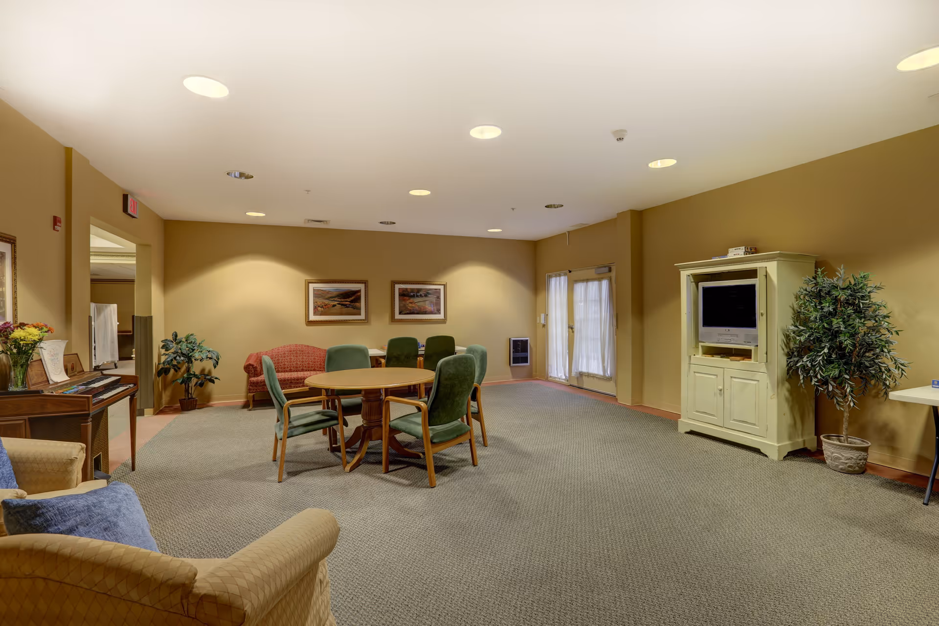 A spacious senior living common room with a round table and chairs, lounge seating, a TV cabinet, plants, and framed artwork on the walls.