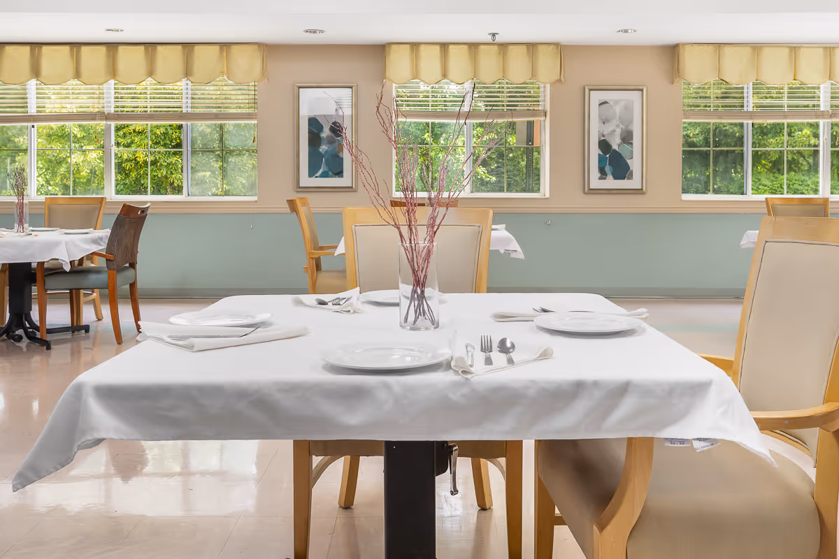 Bright dining room with a table set with a white tablecloth, plates, utensils and a vase of branches, with chairs and large windows showing greenery.