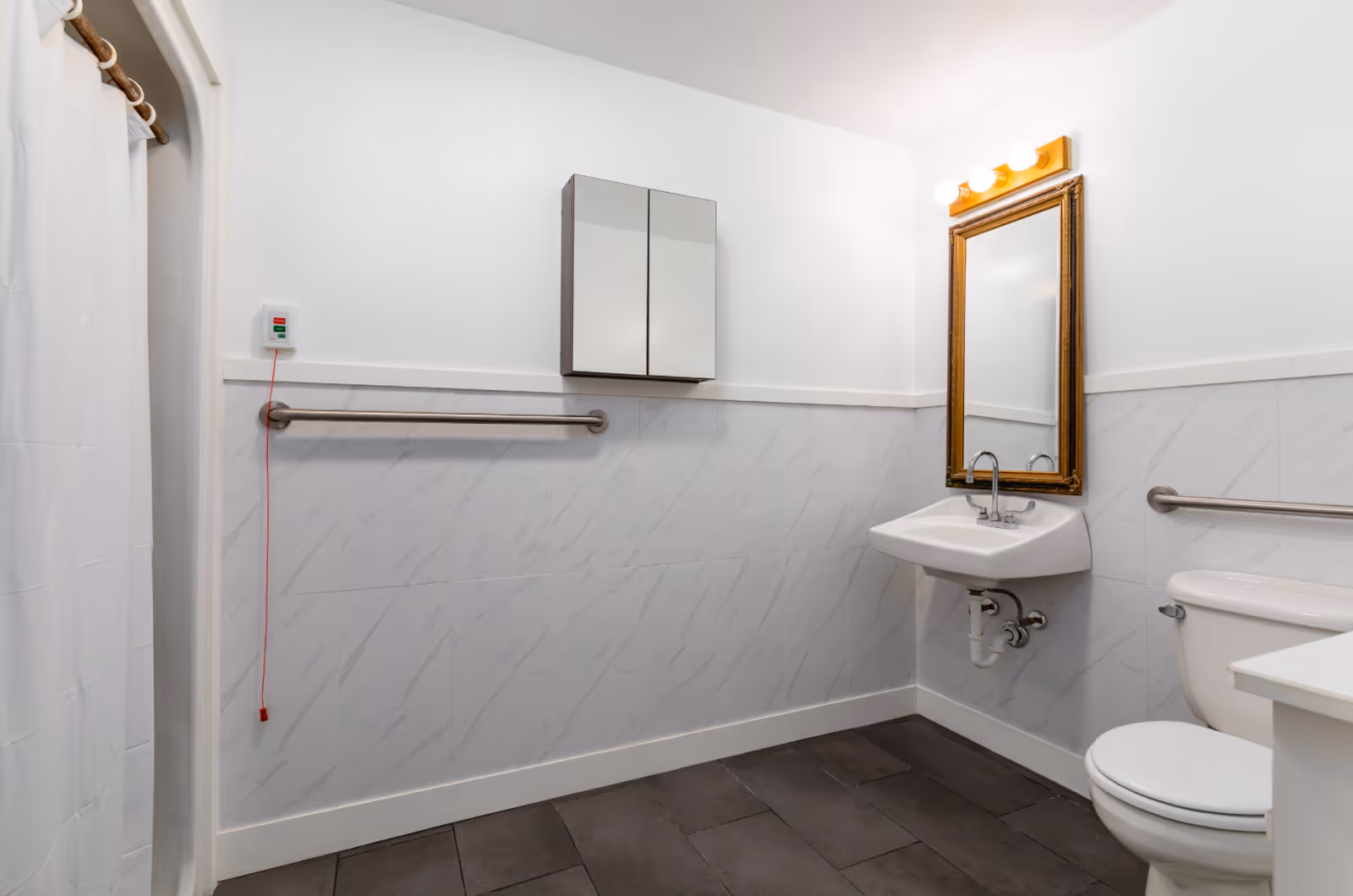 A clean and accessible bathroom with white walls and gray floor tiles. The bathroom features a wall-mounted sink with a gold-framed mirror above it, a toilet with a grab bar on the wall beside it, a shower with a white curtain, and a grab bar on the opposite wall. There is also an emergency pull cord with a red string hanging down.