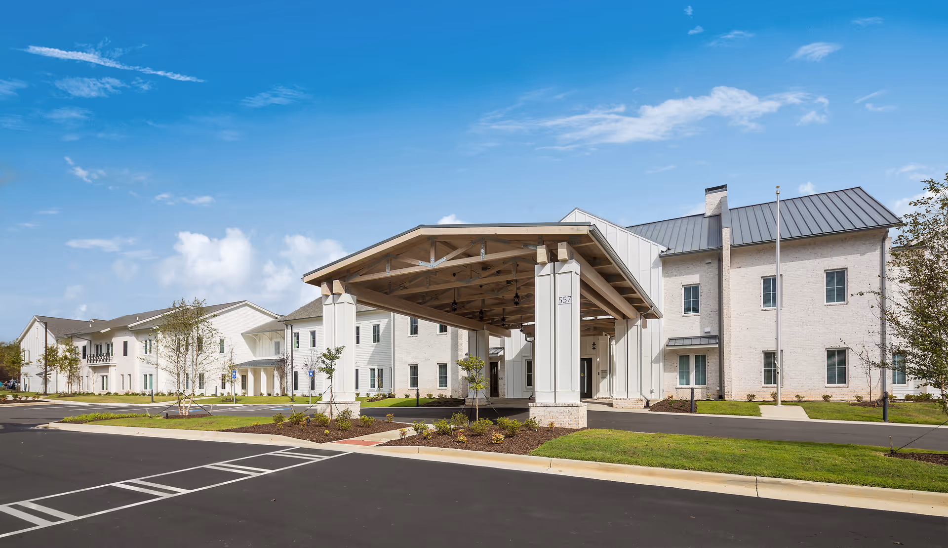 Exterior view of The Claiborne at Westlake senior living facility showing a large covered entrance with white pillars, a paved driveway, and a two-story building with light-colored brick walls and a metal roof under a blue sky with some clouds.