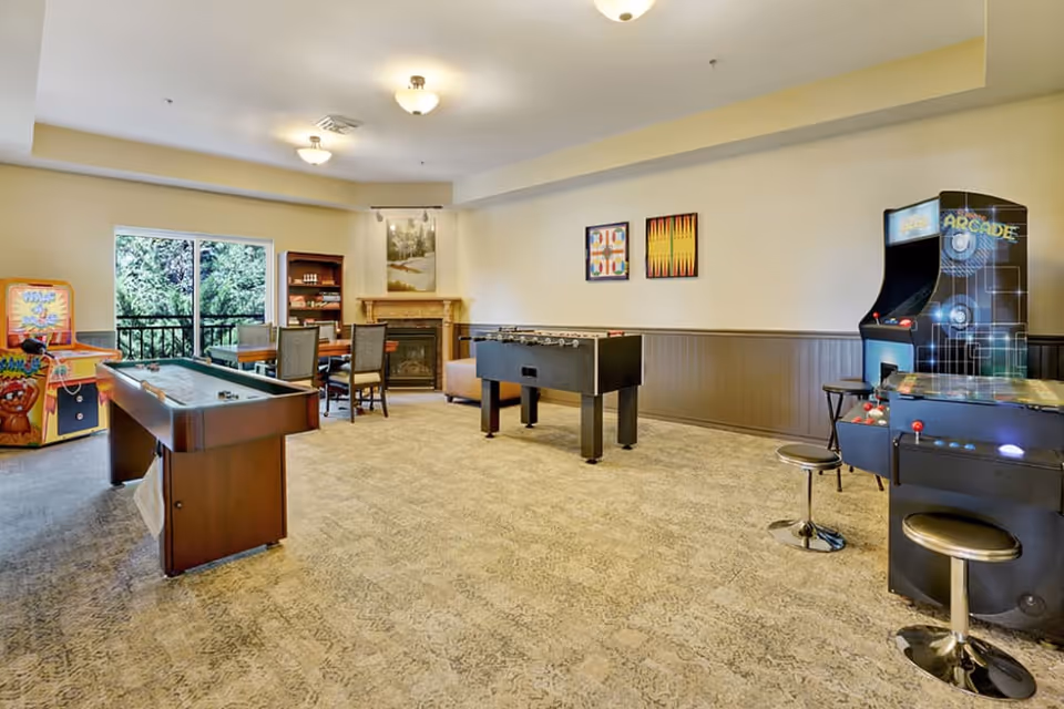 A spacious game room with arcade machines, a foosball table, a shuffleboard table, and a table with chairs near a fireplace. The room has large windows showing greenery outside and framed artwork on the walls.