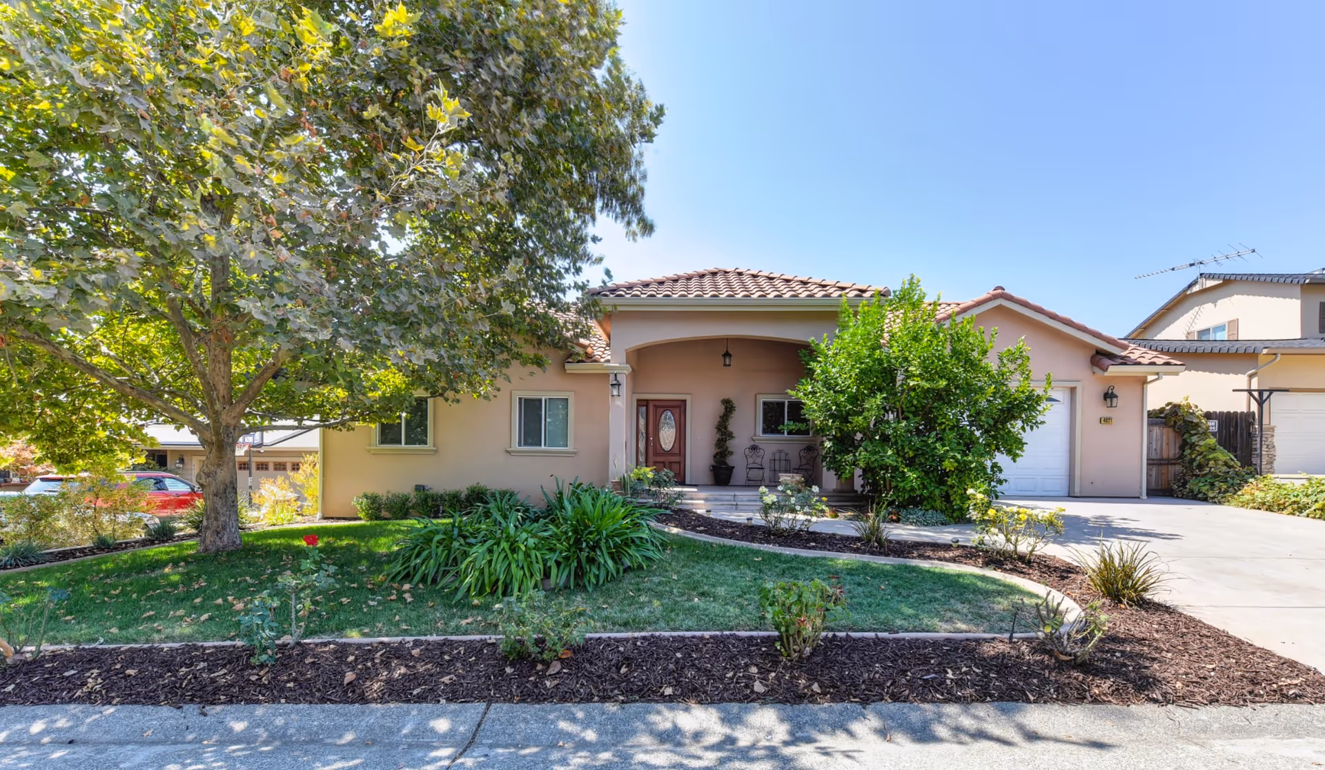 Front exterior view of a single-story house with a tiled roof, a covered porch with two chairs, a wooden front door with glass panel, a large tree and various shrubs and plants in the landscaped front yard, and a driveway leading to a garage.
