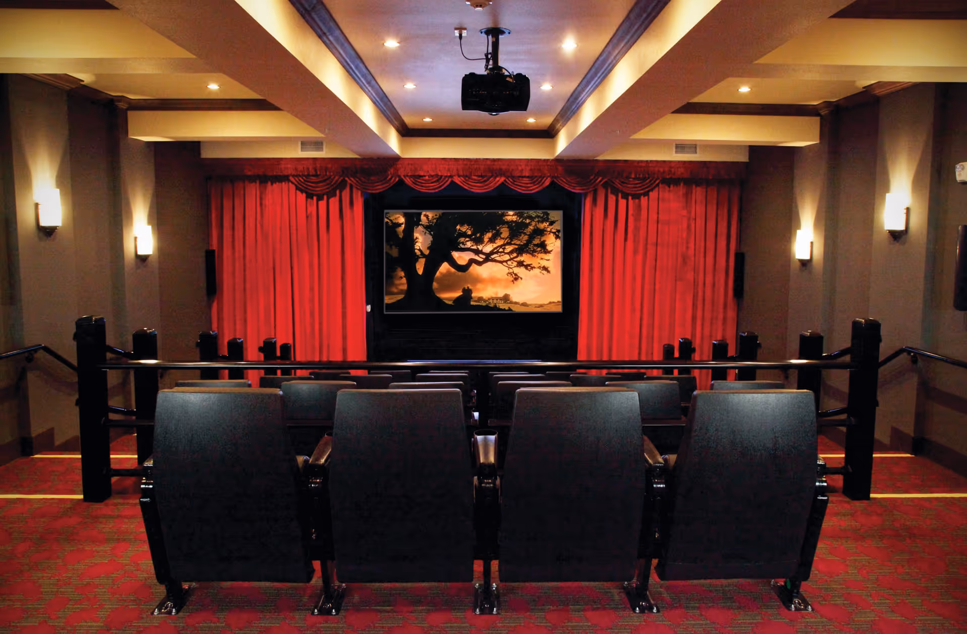 Interior view of a small theater room with red carpet and red curtains surrounding a large screen displaying an image of a tree silhouette at sunset. There are rows of black theater seats facing the screen, and the room is softly lit with wall sconces and ceiling lights.