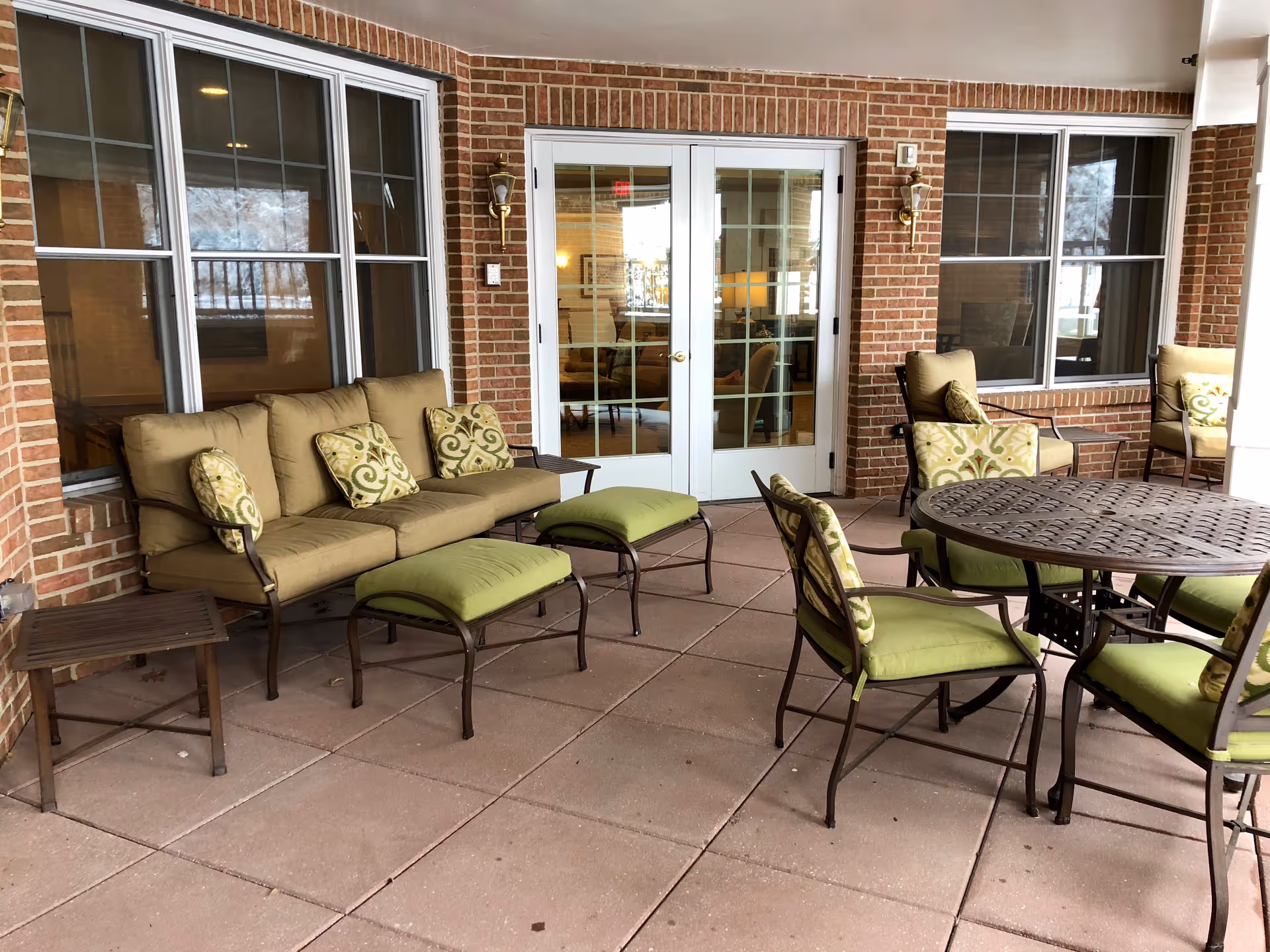 Covered outdoor patio with cushioned seating, green-accented chairs and a round metal table in front of brick walls and glass French doors.