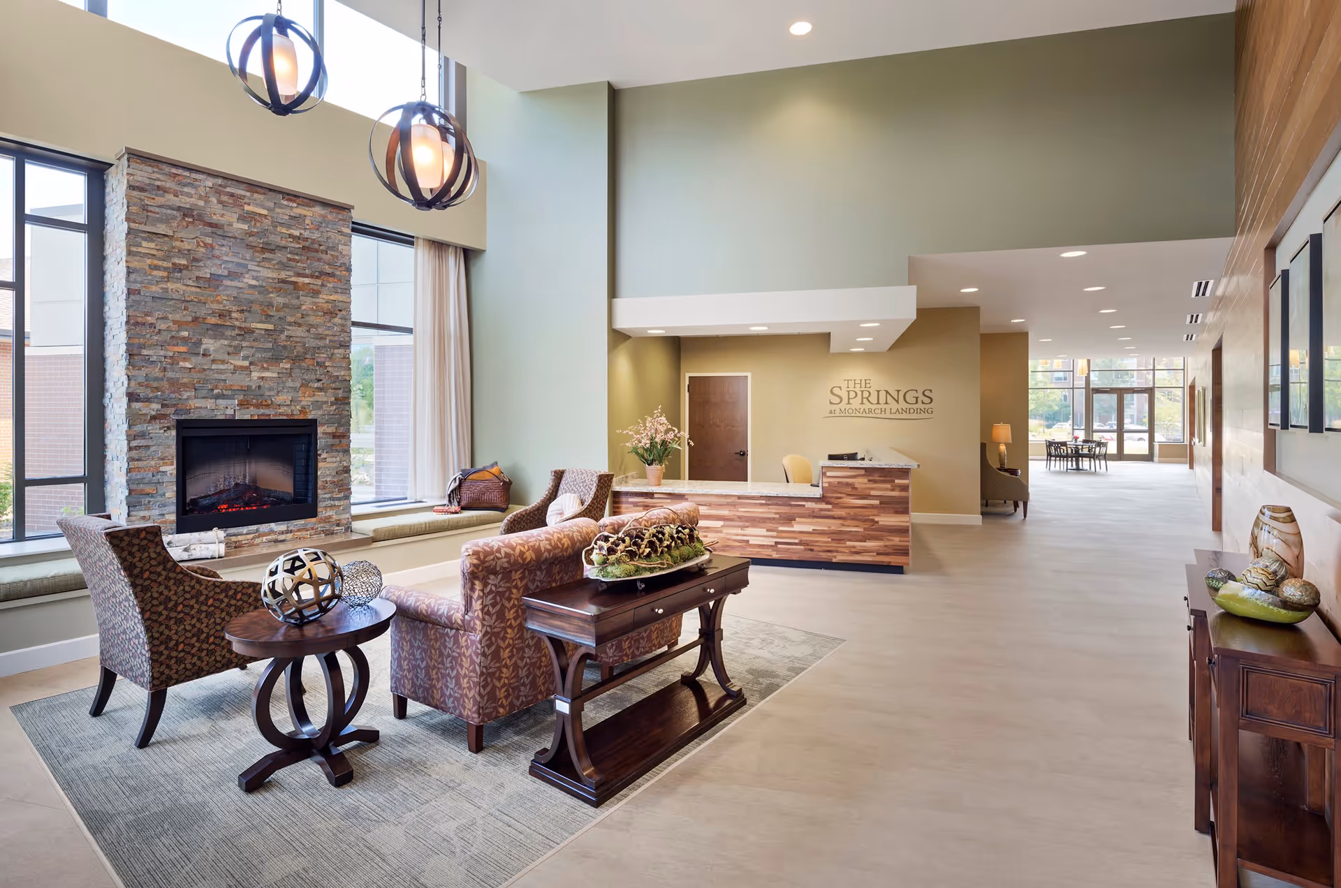 A spacious and well-lit senior living facility lobby with a stone fireplace, comfortable seating including armchairs and a sofa, a wooden coffee table with decorative items, and a reception desk with the sign 'The Springs at Monarch Landing' on the wall behind it. Large windows allow natural light to fill the room, and the hallway extends to a dining area in the background.
