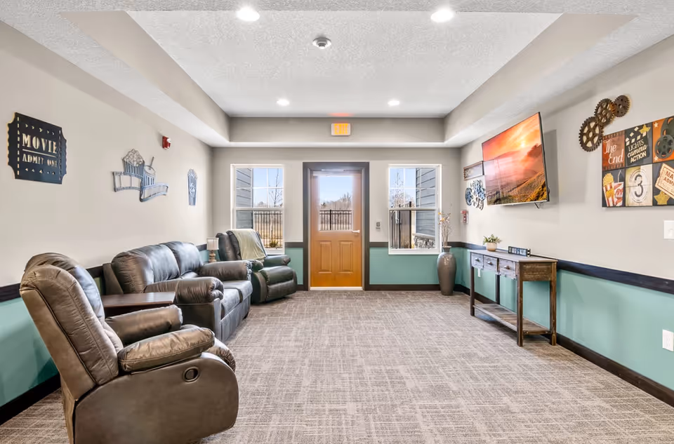 Bright seating lounge with leather recliners facing a wall-mounted TV and a wooden exit door between two windows.