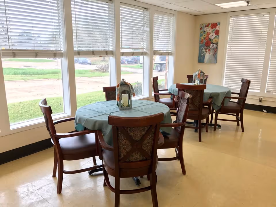 A bright dining area with two round tables covered with light blue tablecloths, each surrounded by four wooden chairs with patterned backs. Each table has a decorative lantern centerpiece. Large windows with white blinds allow natural light to fill the room, and a colorful floral painting hangs on the wall.