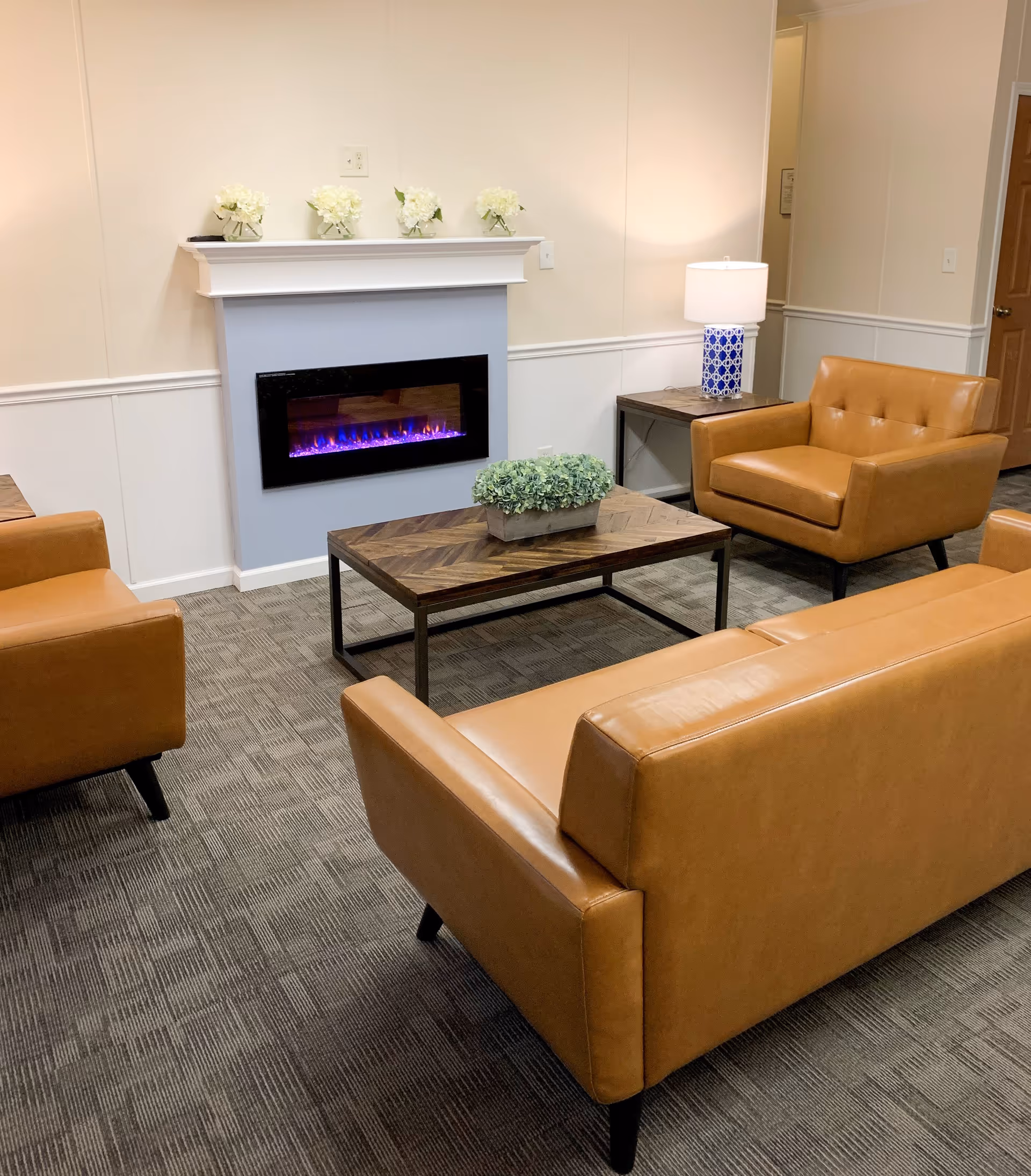Cozy lounge with tan leather sofas and armchairs arranged around a wooden coffee table in front of a wall-mounted electric fireplace.