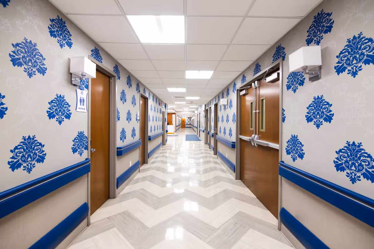 Long interior hallway in a nursing facility with blue damask wallpaper, handrails, chevron tile floor, and multiple wooden doors.