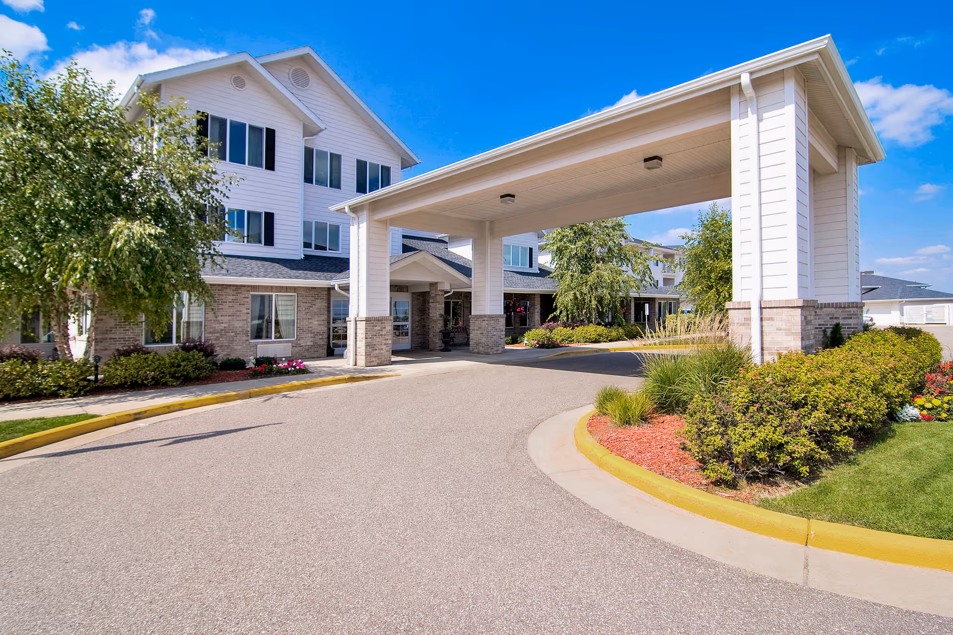Exterior view of Oakwood Hills - Sky Active Living facility showing a covered driveway entrance with white pillars and a multi-story building in the background under a blue sky with some clouds. The area is landscaped with green bushes, trees, and colorful flowers.