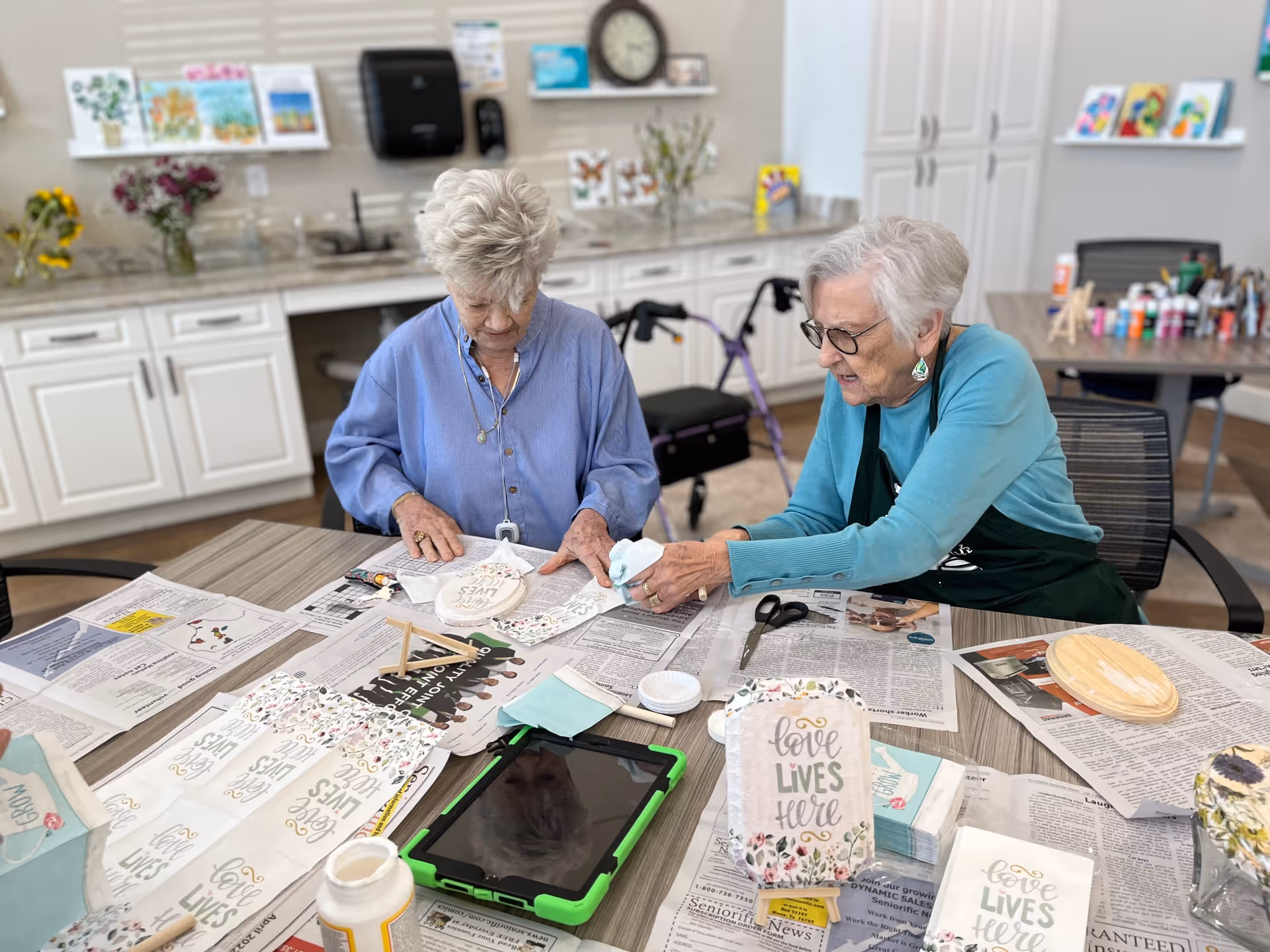 Two elderly women sitting at a table in a craft room, working on decorative plaques with the phrase 'love lives here'. The table is covered with newspapers, craft supplies, and an electronic tablet. The background shows a countertop with cabinets, flowers, and art supplies.