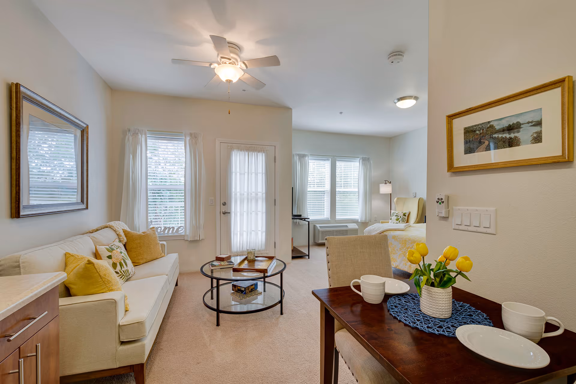 A bright and cozy living space in a senior living facility featuring a beige sofa with yellow and floral cushions, a round glass coffee table, a dining table set with white dishes and a vase of yellow tulips, and a bed near large windows with white curtains. The room has light-colored walls, carpeted floor, ceiling fan, and framed artwork on the walls.