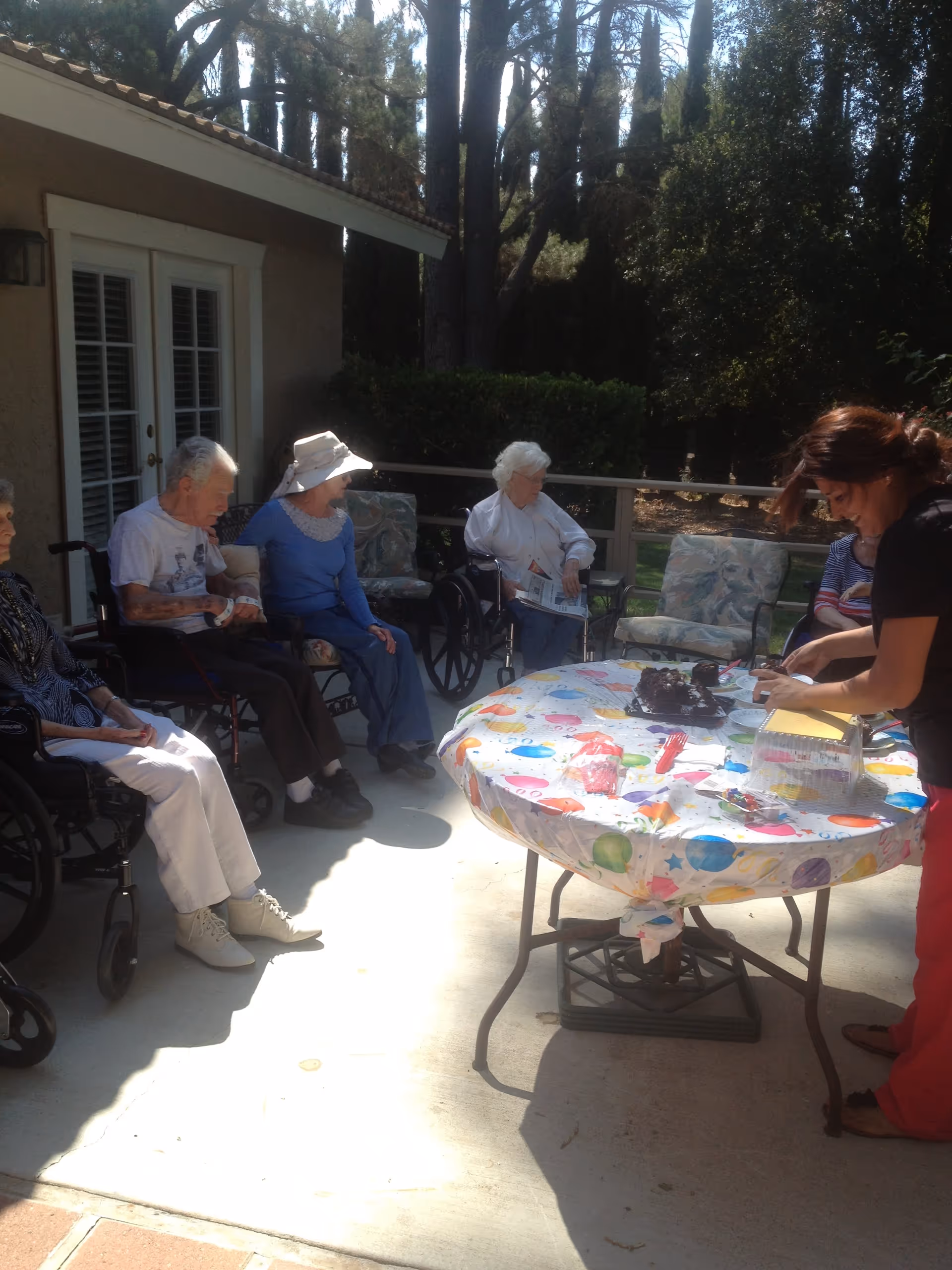 A group of elderly people sitting outdoors on a patio, some in wheelchairs, with a woman standing at a table covered with a colorful tablecloth. The table has a cake and some party supplies on it. The setting is surrounded by trees and bushes.