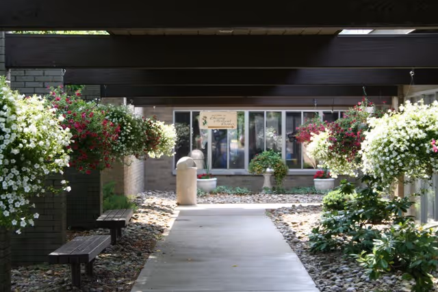 A covered outdoor walkway with hanging flower baskets on both sides, benches along the left side, and a pathway leading to a building entrance with large windows and a sign above the door.