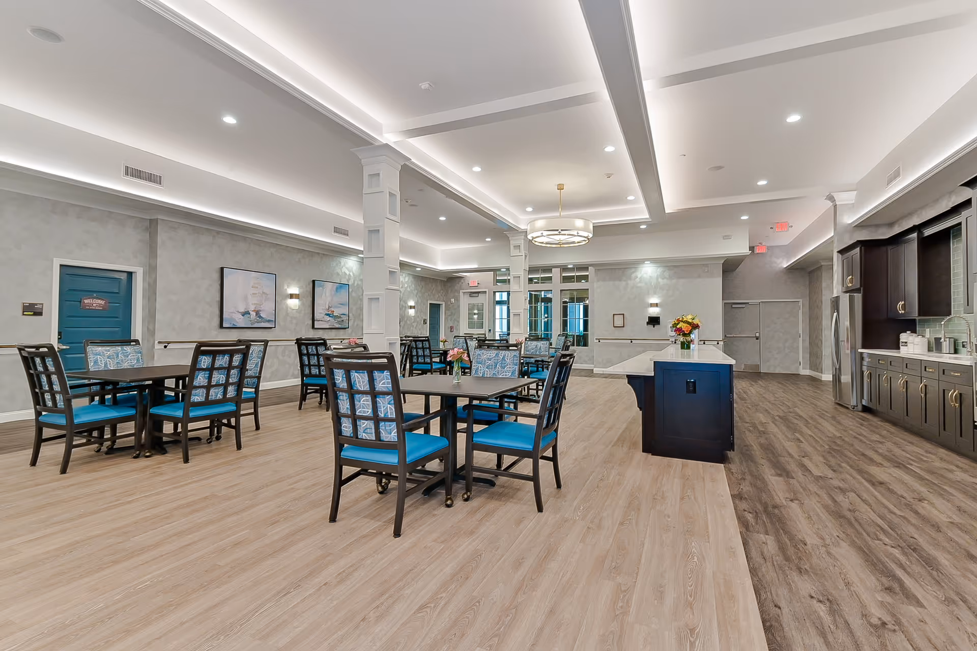 A spacious and well-lit dining area in a senior living facility featuring multiple tables with blue cushioned chairs. The room has light wood flooring, decorative wall art, and a modern kitchen area with dark cabinetry and stainless steel appliances on the right side. The ceiling has recessed lighting and a large circular chandelier.