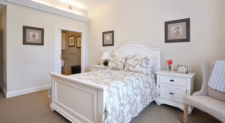 A bright bedroom with a white wooden bed frame and floral patterned bedding. There are two matching white nightstands on either side of the bed, one with a lamp and the other with a small vase of red flowers and a picture frame. A beige upholstered chair with a light blue throw blanket is positioned to the right. The walls are decorated with framed shell artwork, and an open doorway leads to a bathroom.