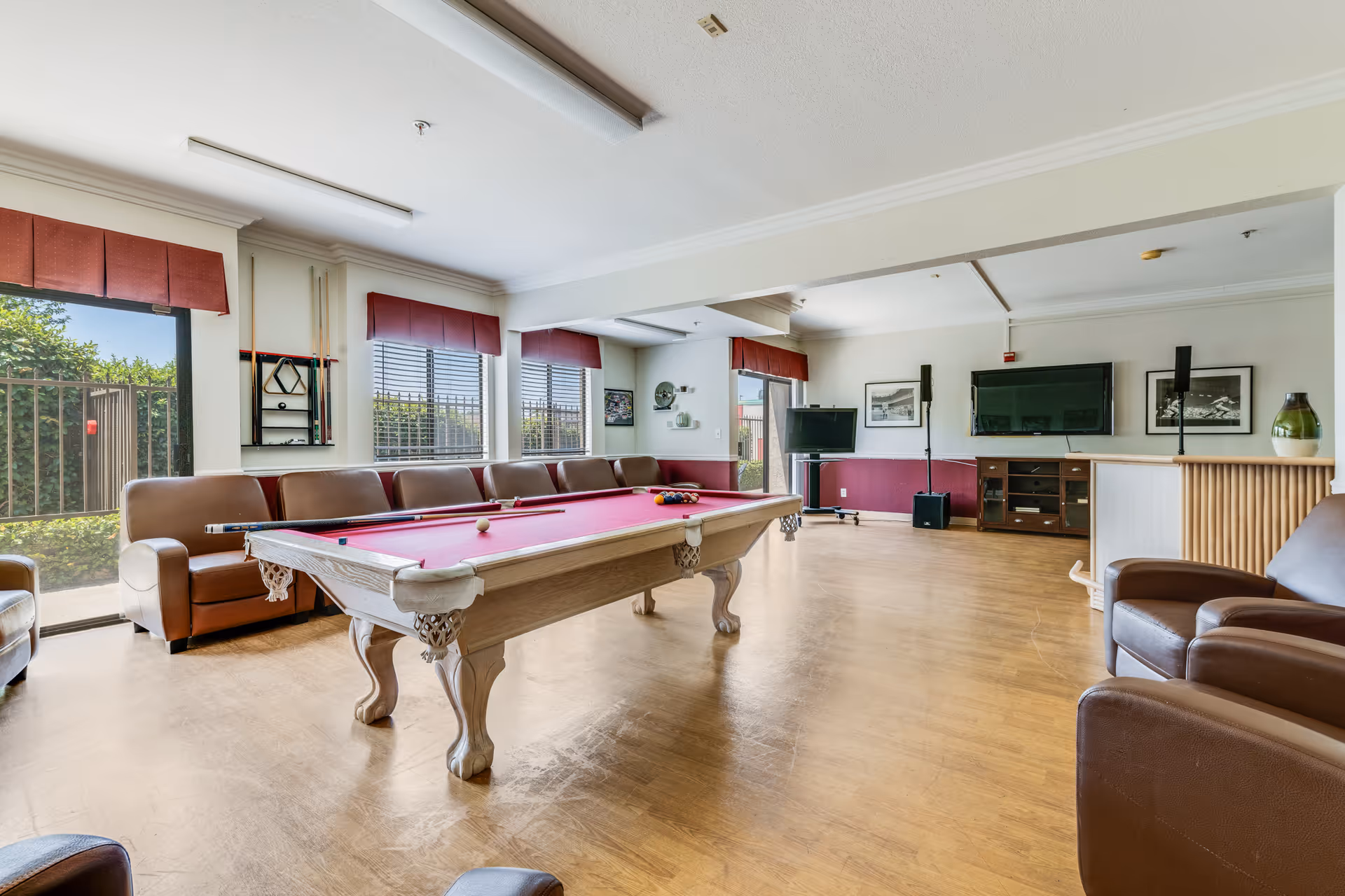 A spacious recreational room with a red felt pool table in the center, surrounded by brown leather sofas and armchairs. The room has large windows with red valances letting in natural light, a TV mounted on the wall, and a wooden cabinet beneath it. The floor is wooden, and there are pool cues mounted on the wall near the windows.