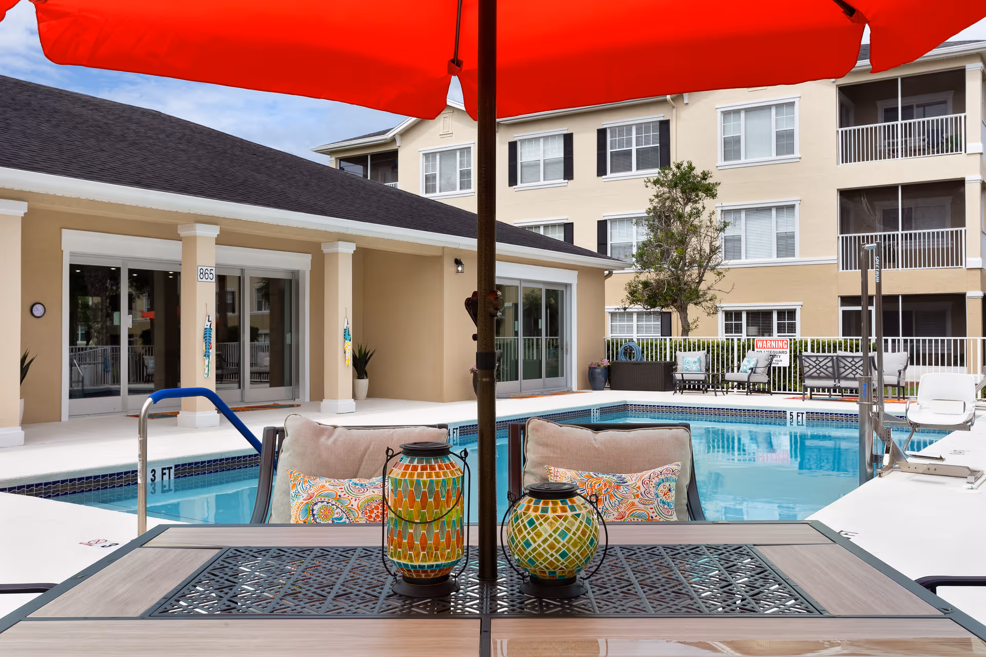 Outdoor pool area at Glenbrooke Senior Living with a table and chairs under a large red umbrella in the foreground. The pool is surrounded by a beige building with sliding glass doors and a multi-story residential building in the background. There are colorful cushions on the chairs and decorative lanterns on the table.
