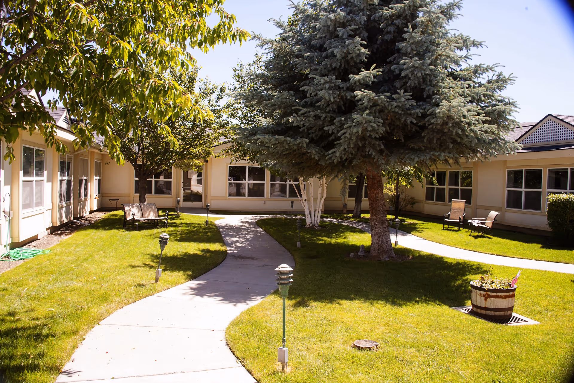 Outdoor courtyard area at Settler's Park Senior Living with a winding concrete pathway, green grass, several trees including a large pine tree, and outdoor seating with chairs and benches near the building windows.